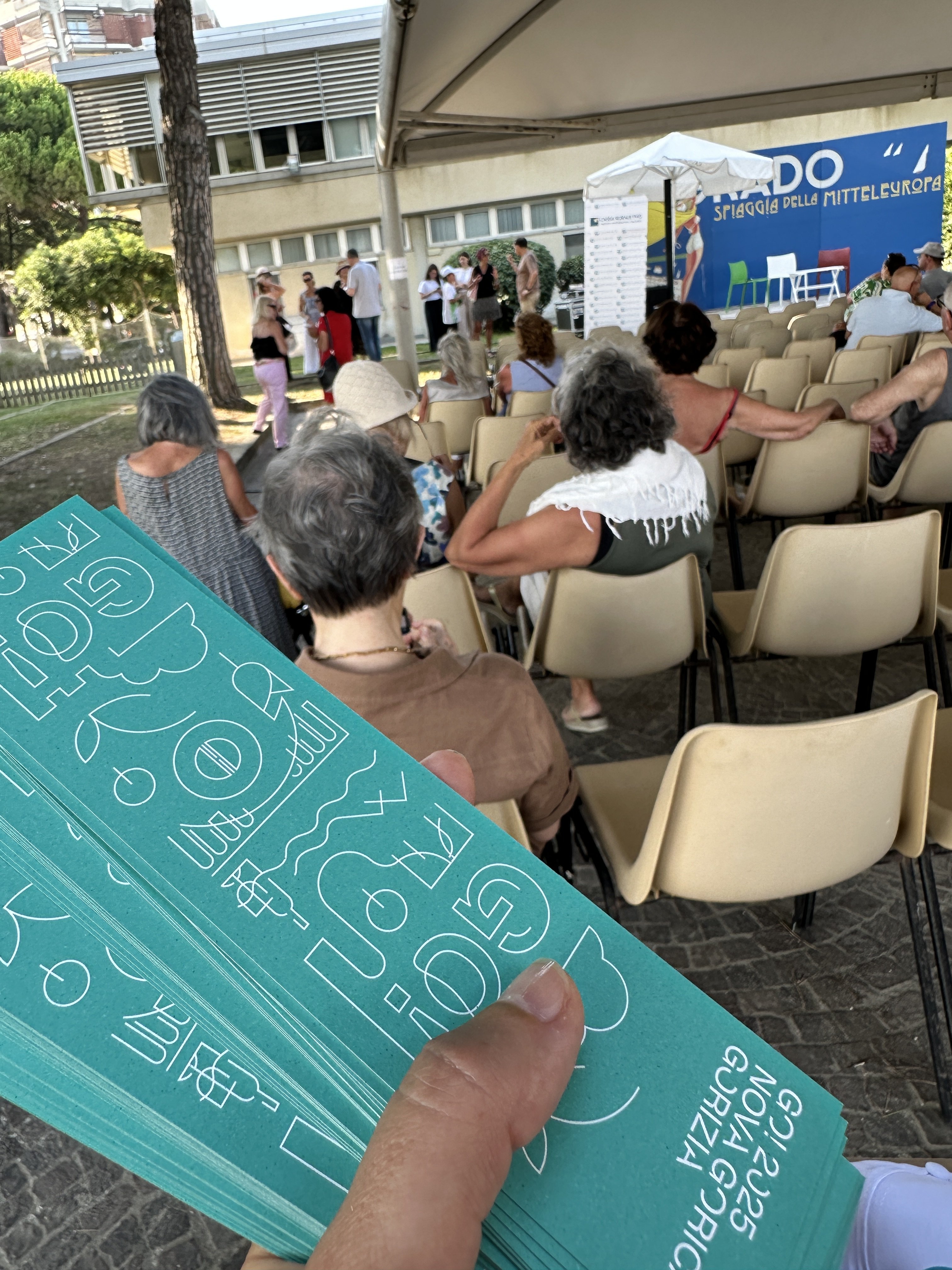 GO! 2025 all’evento Grado Spiaggia della Mitteleuropa GO! 2025 all'evento Grado Spiaggia della Mitteleuropa. Una mano regge del materiale promozionale mentre si svolge la presentazione dell'evento all'aperto.