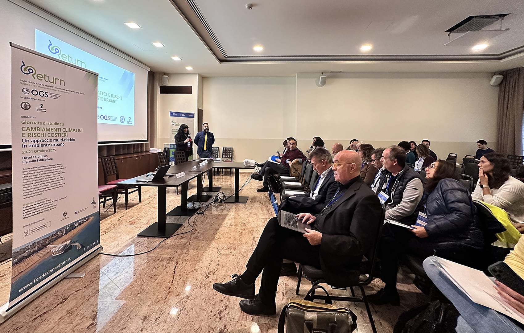 Sala conferenze durante l’apertura dell’evento RETURN a Lignano Veduta laterale della sala conferenze dell’evento “Cambiamenti climatici e rischi costieri” a Lignano, con partecipanti seduti e relatori pronti a iniziare.